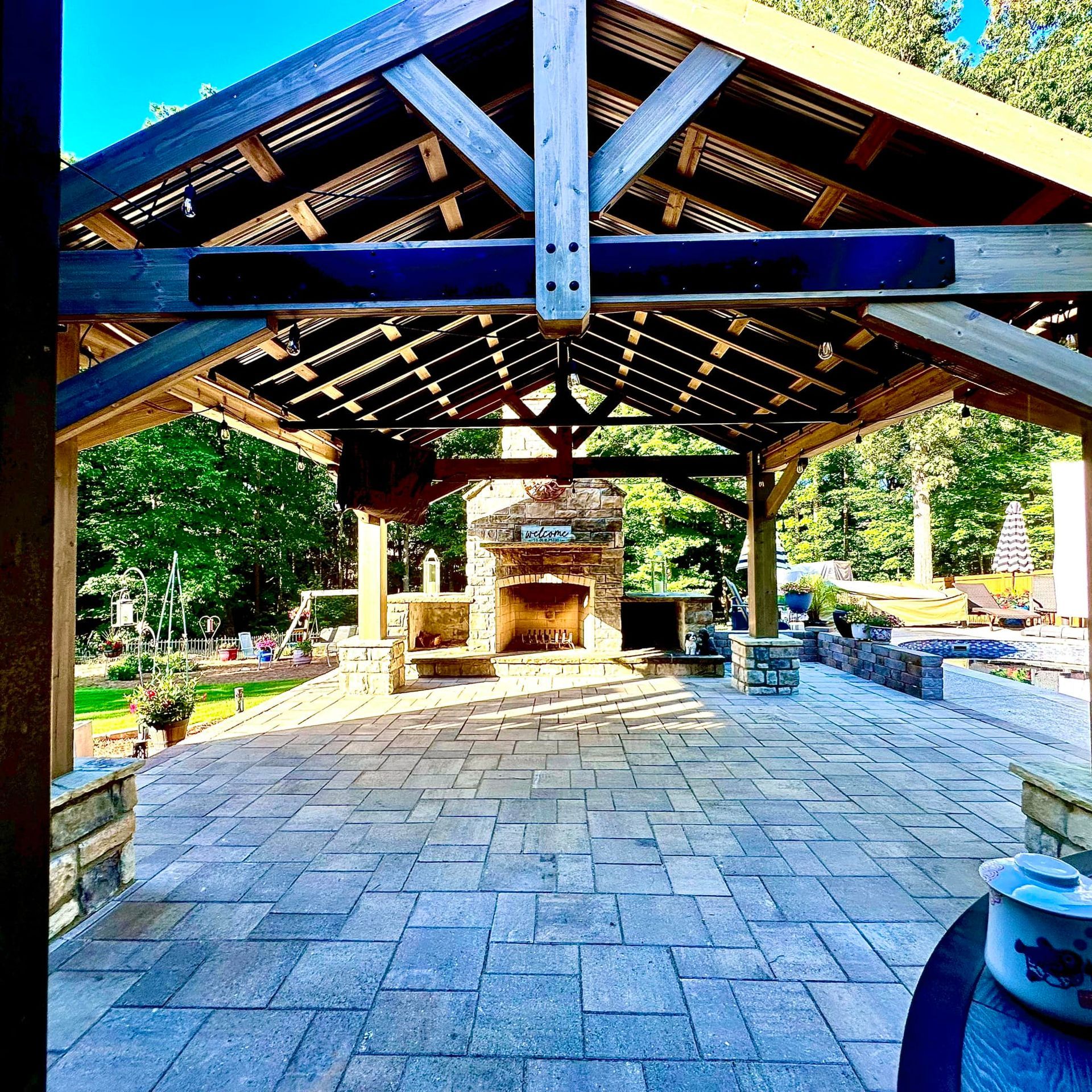 Open-air stone patio with a wooden pavilion and outdoor fireplace, surrounded by greenery.