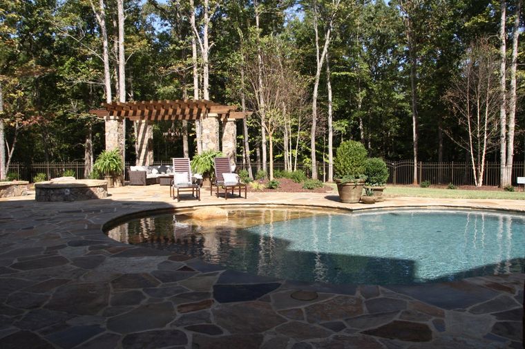 Backyard pool with stone patio, pergola, lounge chairs, and trees in the background