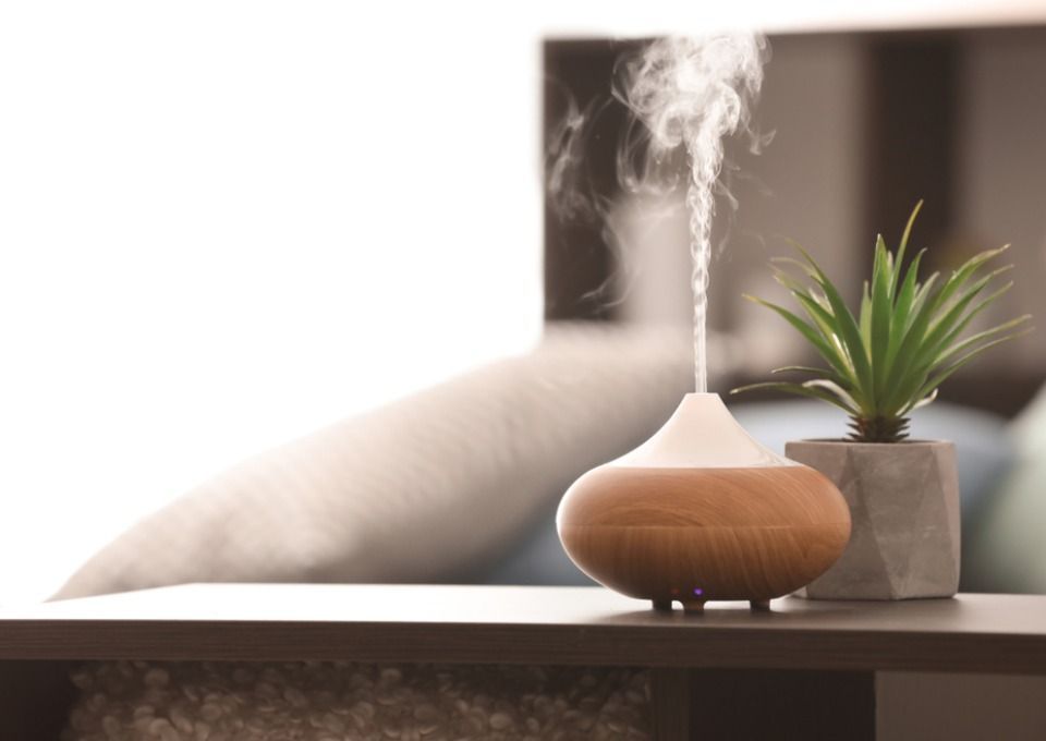 A wooden oil diffuser releasing a stream of mist next to a small potted succulent on a wooden surface.