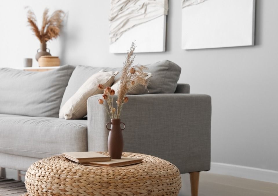 A gray sofa with a woven ottoman, dried floral arrangements, and abstract art in a minimalist, neutral-toned living room.