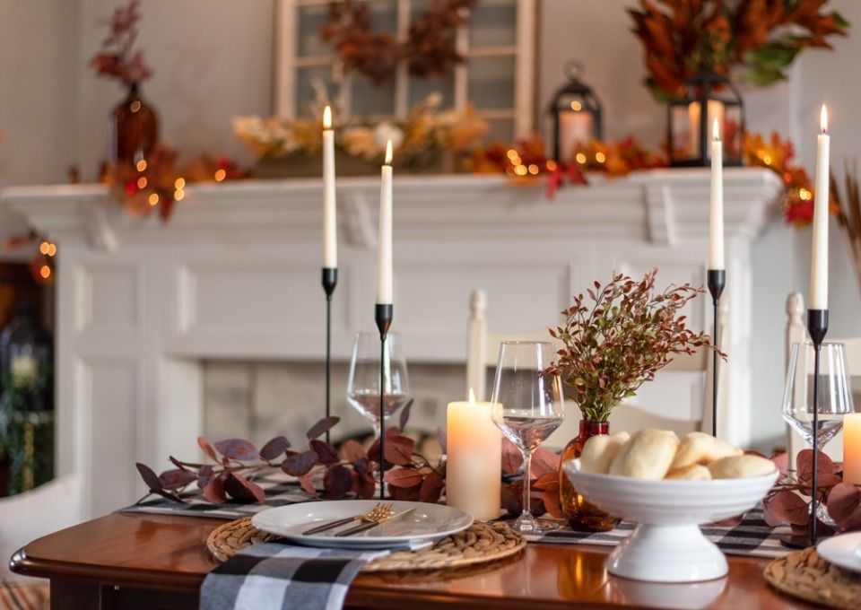 A festive Thanksgiving dining table set with white plates, candles, autumn foliage, and a bowl of rolls by a fireplace.