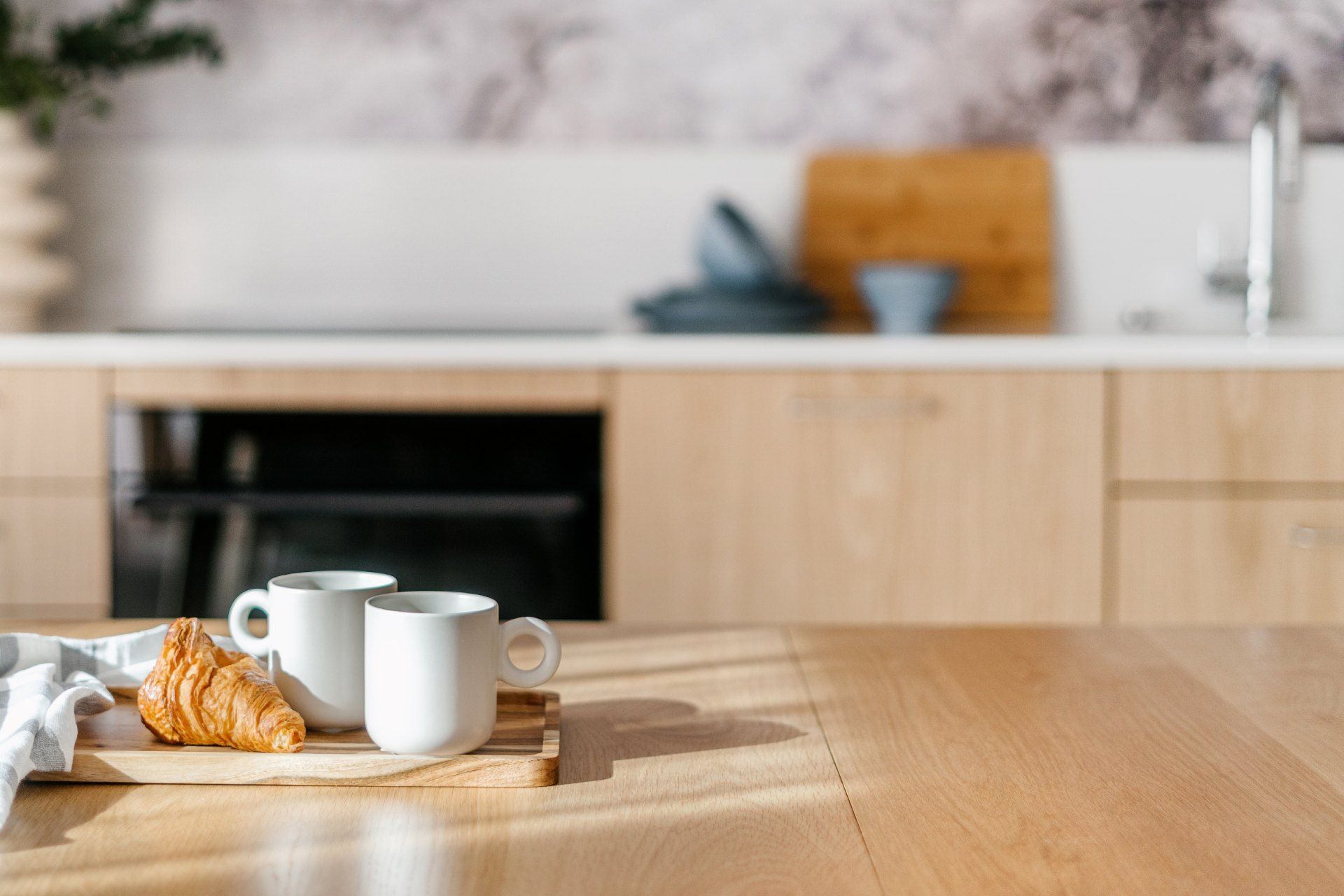 A croissant and two white mugs on a wooden board on a kitchen table, with a blurred modern kitchen in the background.