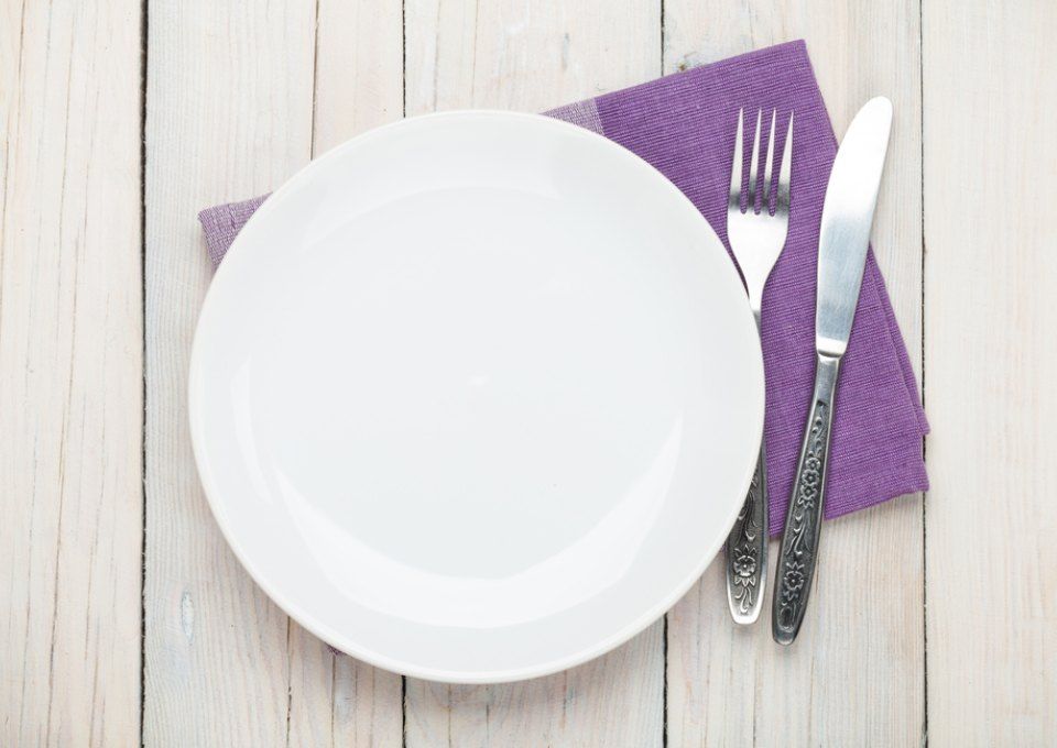 An empty white plate, a fork, and a knife placed on a purple napkin over a light-colored wooden table.