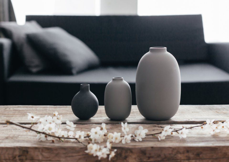 Three minimalist grey vases of varying sizes on a rustic wooden table with white blossoms, in front of a dark sofa.