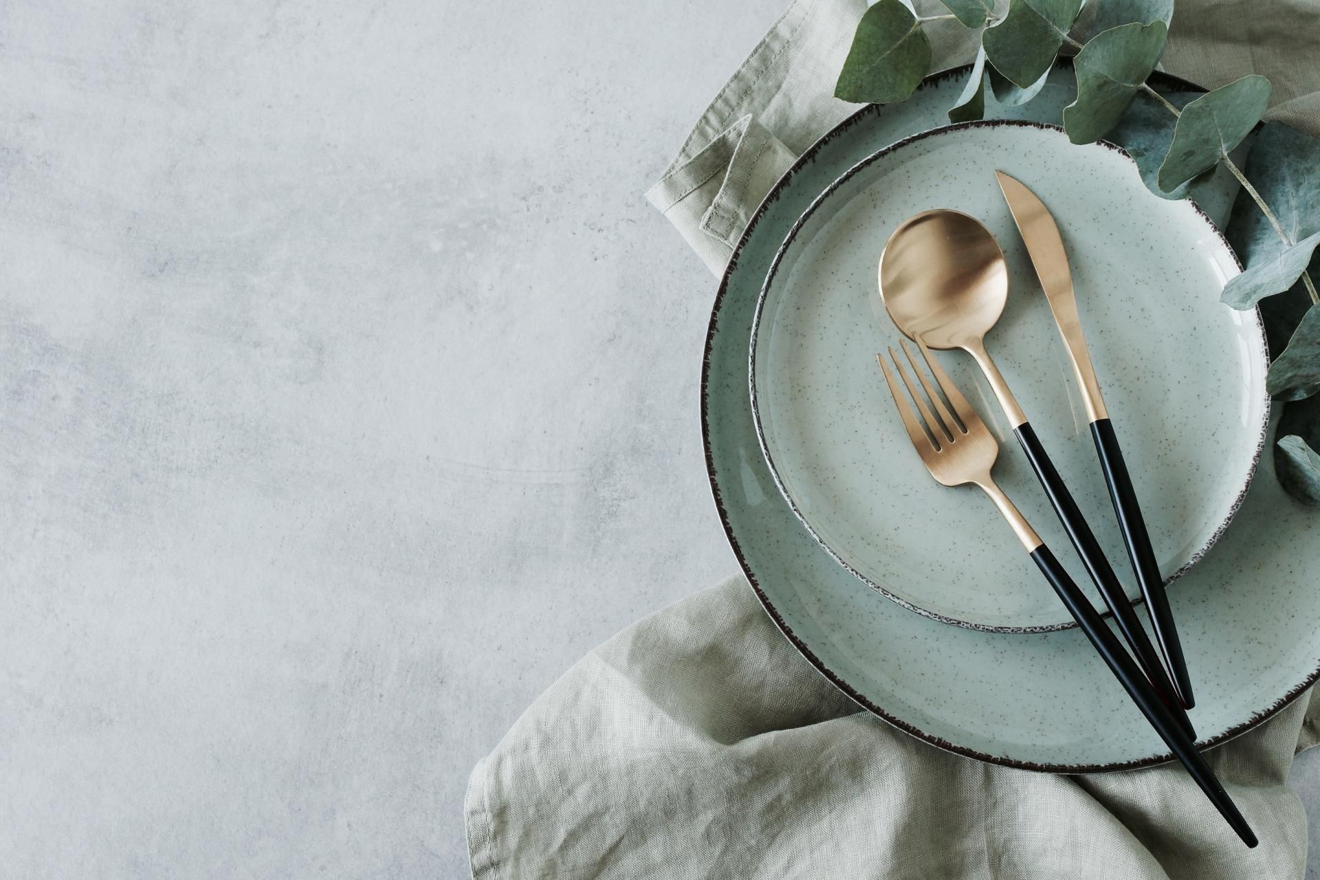 A place setting with two grey plates, a gold fork, spoon, and knife, and a linen napkin on a textured grey background.