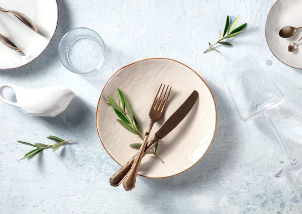 A minimalist table setting with plates, bronze cutlery, a glass, a small white creamer, and olive sprigs on blue stone.