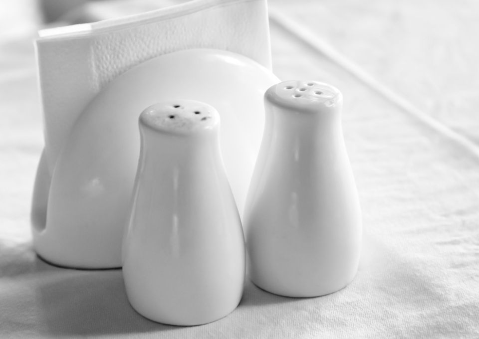 Two white ceramic salt and pepper shakers in front of a white napkin holder on a table.