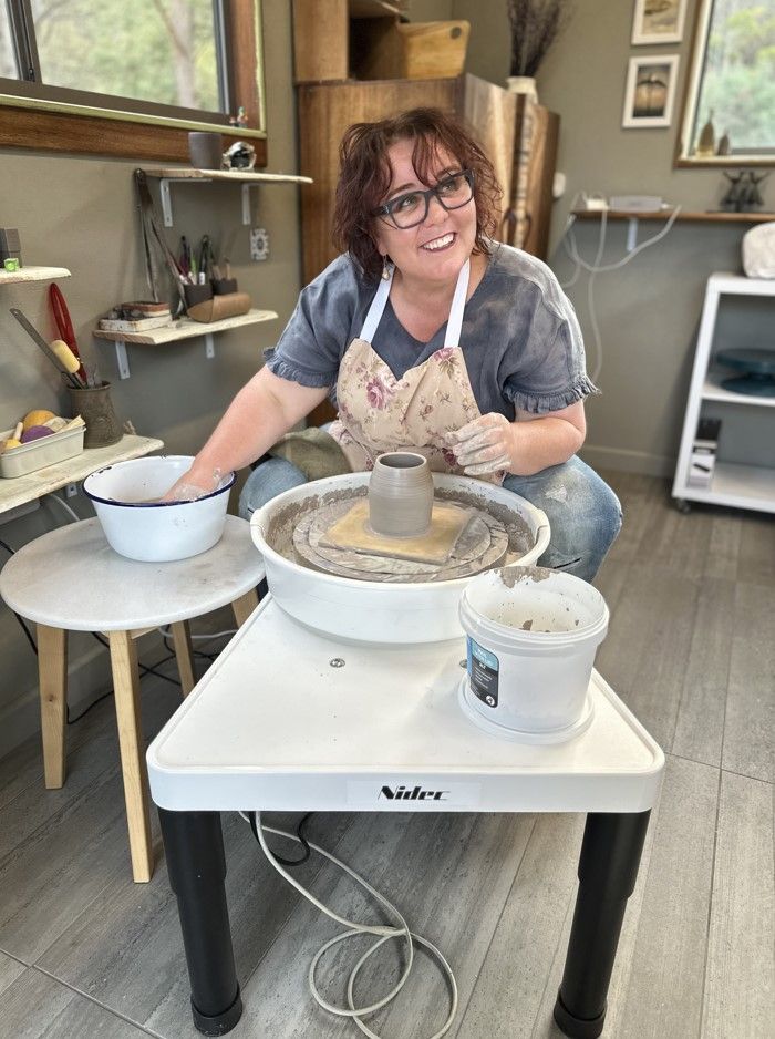 A person wearing an apron works at a pottery wheel in a studio, smiling while shaping a clay vessel.