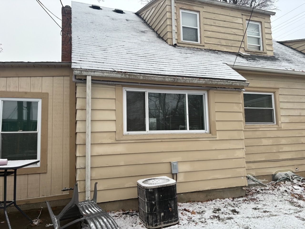 A tan, two-story house with a light dusting of snow on the roof and ground, featuring a central window and AC unit.
