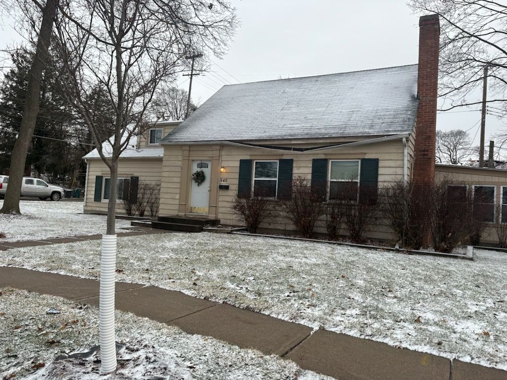 A light-colored house with dark shutters and a brick chimney sits on a snowy lawn on an overcast day.