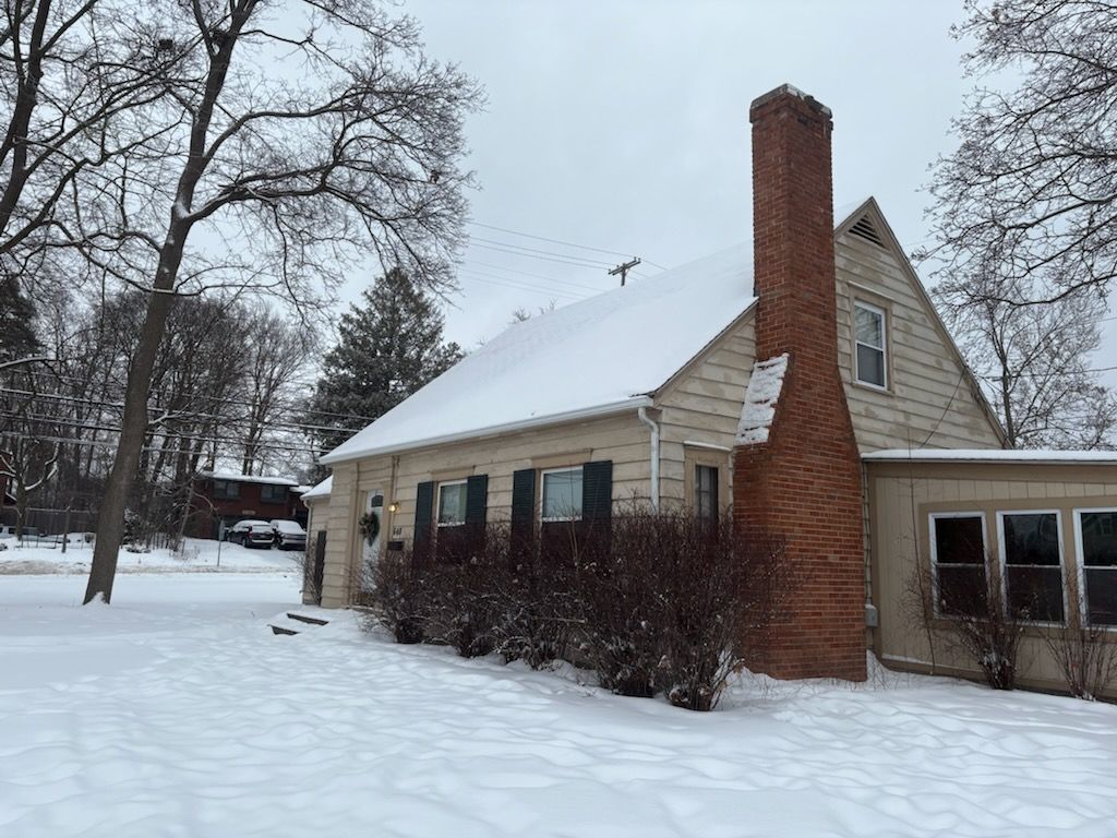 A tan, one-story house with a brick chimney and dark shutters, surrounded by snow on a cloudy winter day.