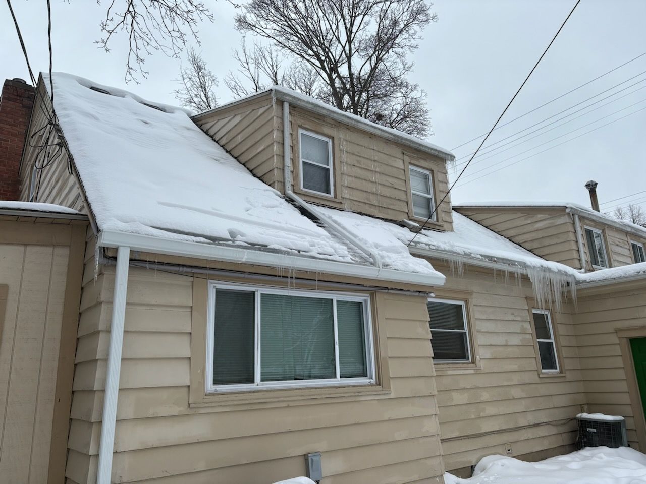 A tan house with beige siding and windows sits under a snow-covered roof, with icicles hanging from the gutters.