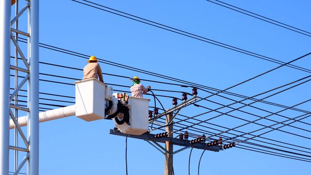 Operai di una società di servizi pubblici riparano le linee elettriche aeree su una piattaforma elevatrice sotto un cielo azzurro.