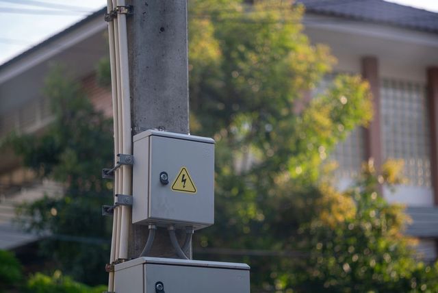 Scatola elettrica grigia con cartello di avvertimento montata su un palo della luce all'aperto, con case sfocate sullo sfondo.