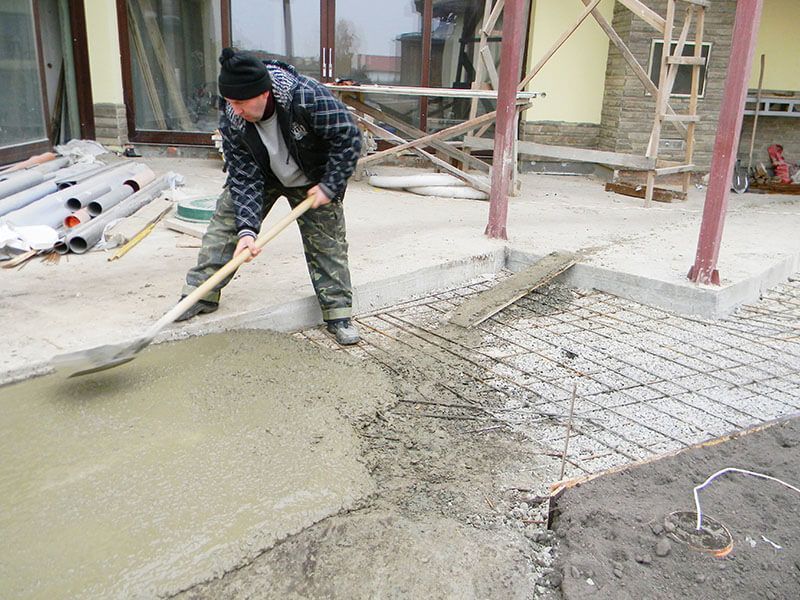 Man spreading wet concrete with a shovel on a patio, near a building and construction materials.