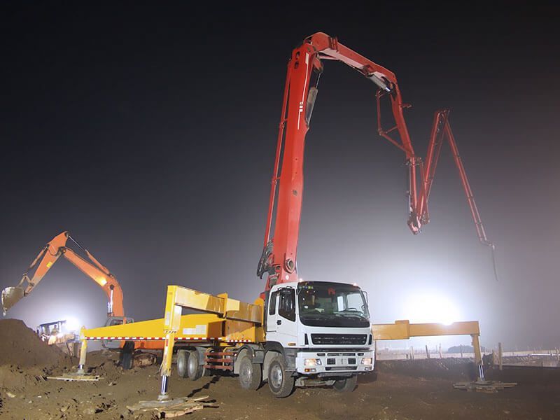 A large red concrete pump truck is working at night, with an excavator in the background.