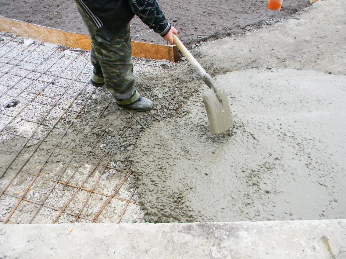 Person using a shovel to spread wet concrete over metal rebar during construction.