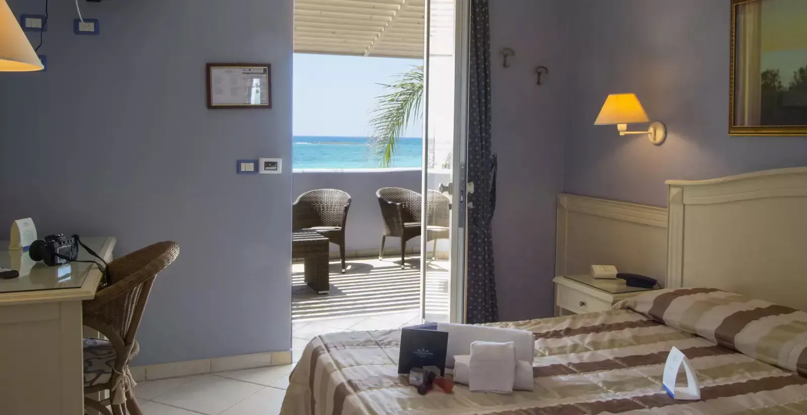 Hotel room with a bed, desk, and open door to a balcony overlooking the sea.