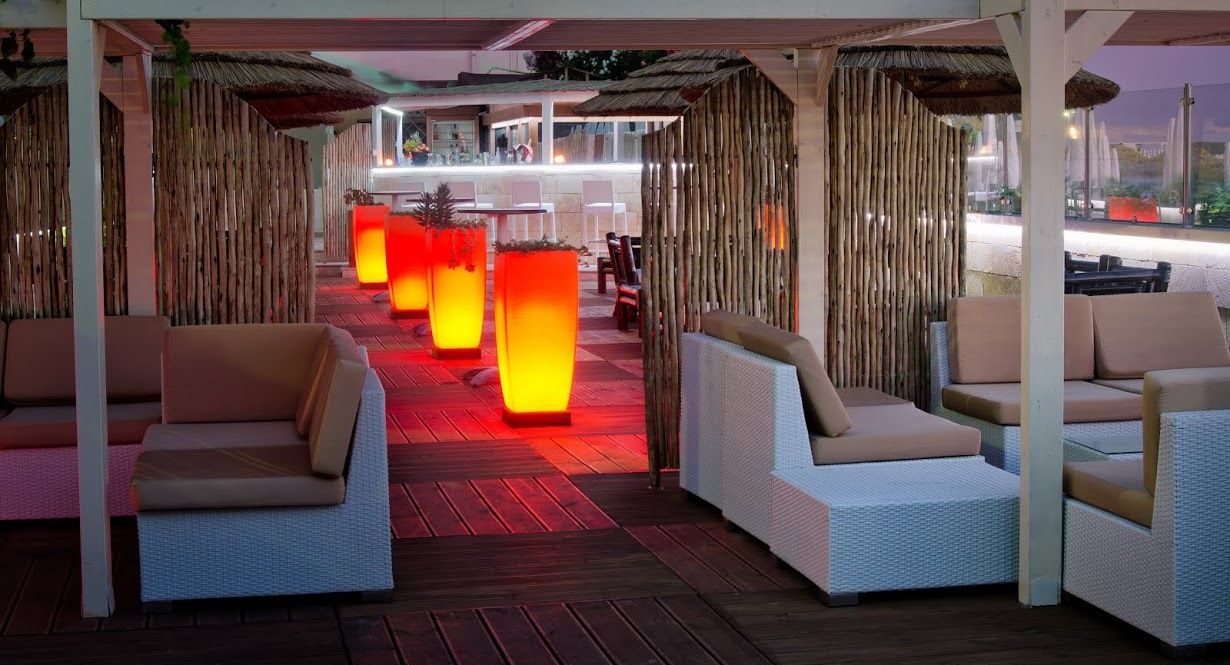 Outdoor lounge with white wicker furniture, red-lit pillars, bamboo walls, wooden floor, and thatched roof.
