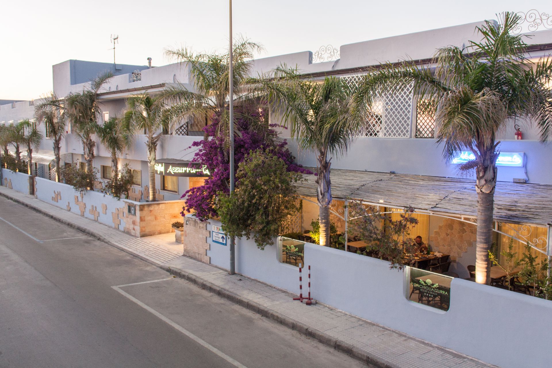 Edificio bianco con palme e fiori viola accanto a una strada al tramonto.