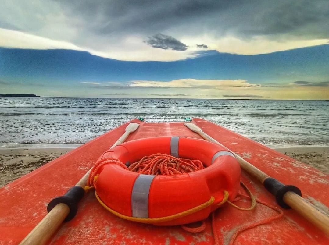 Barca a remi rossa su una spiaggia sabbiosa con salvagente e remi, rivolta verso il mare sotto un cielo nuvoloso.