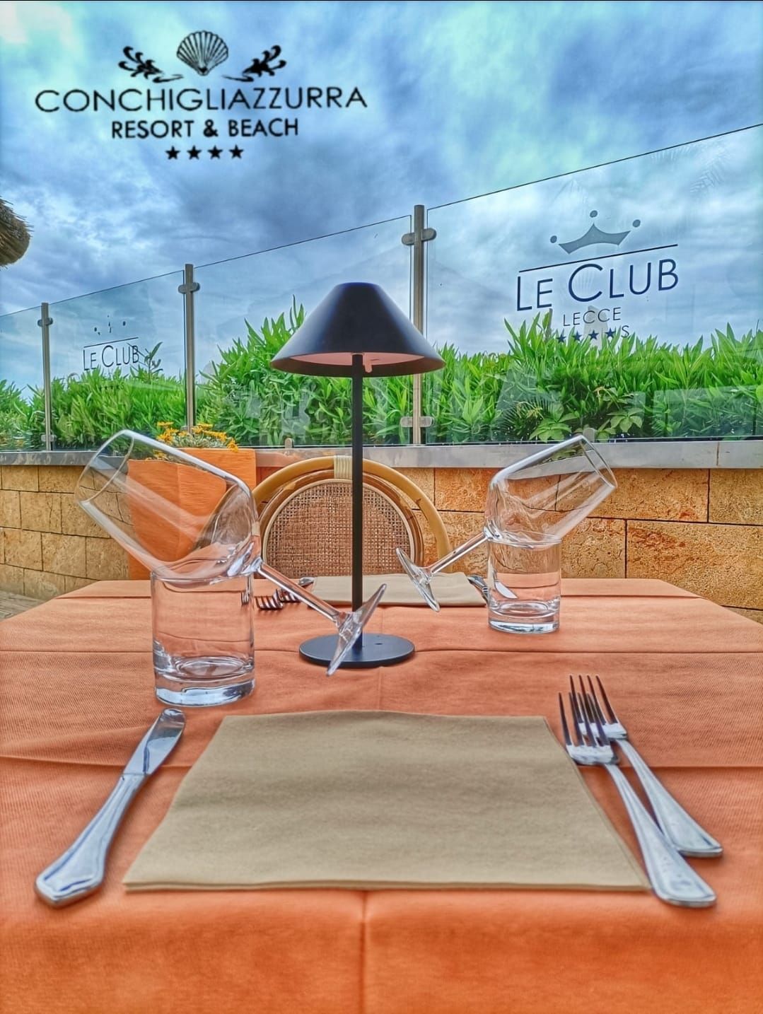 Tavolo da ristorante apparecchiato per cenare, con vista sul verde e sul cielo nuvoloso.