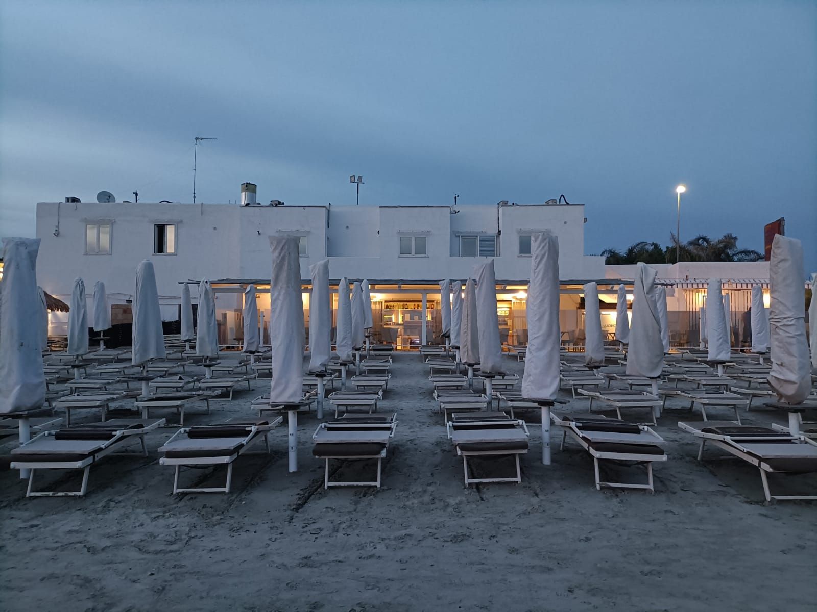 Spiaggia con file di ombrelloni chiusi e sedie a sdraio, di fronte alla quale si erge un edificio bianco al tramonto.