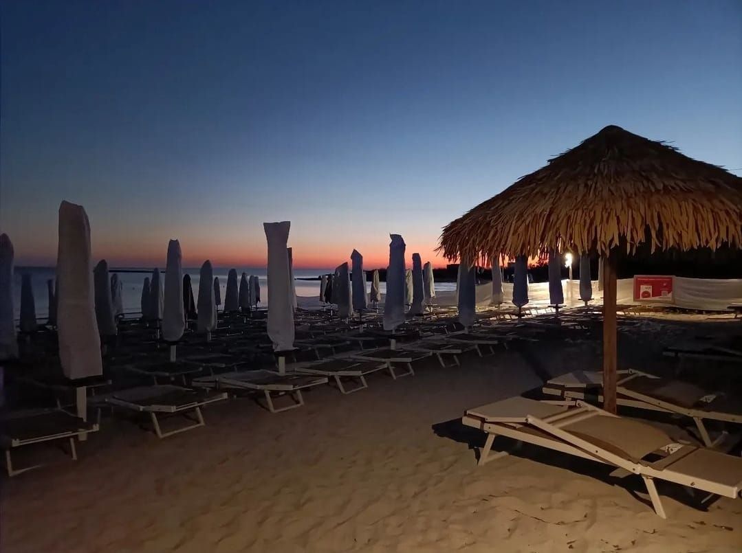 Scena di spiaggia al tramonto con ombrelloni chiusi, sedie a sdraio e un ombrellone di paglia.