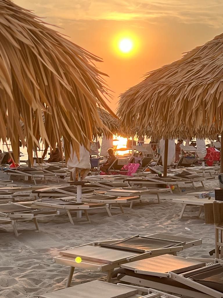 Scena di spiaggia al tramonto con ombrelloni di paglia, sedie a sdraio e persone.