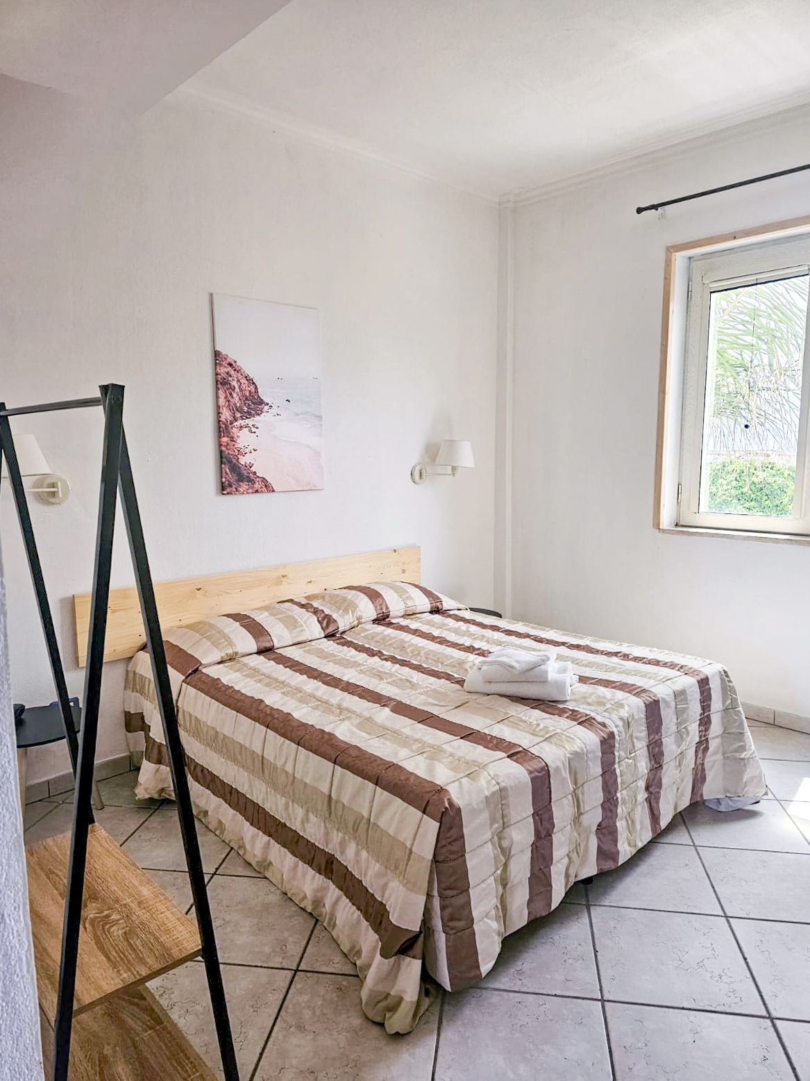 Camera da letto con letto matrimoniale, piumone a righe, pareti bianche, finestra, quadri e appendiabiti.