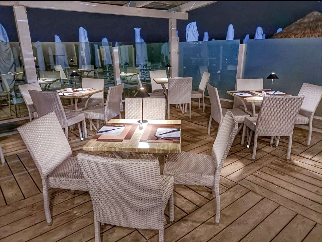 Posti a sedere all'aperto del ristorante con sedie in vimini, tavoli apparecchiati con tovagliette, spiaggia sullo sfondo.