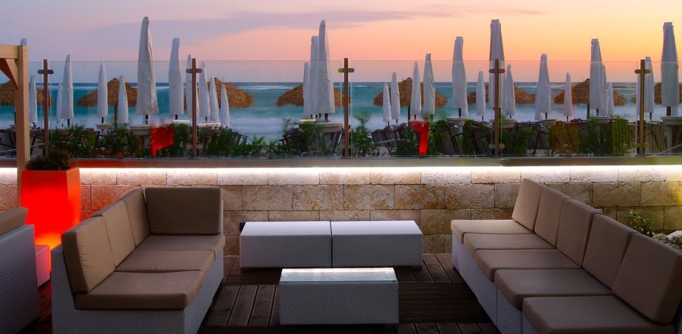 Area lounge all'aperto con divani e tavoli con vista sugli ombrelloni e sull'oceano al tramonto.