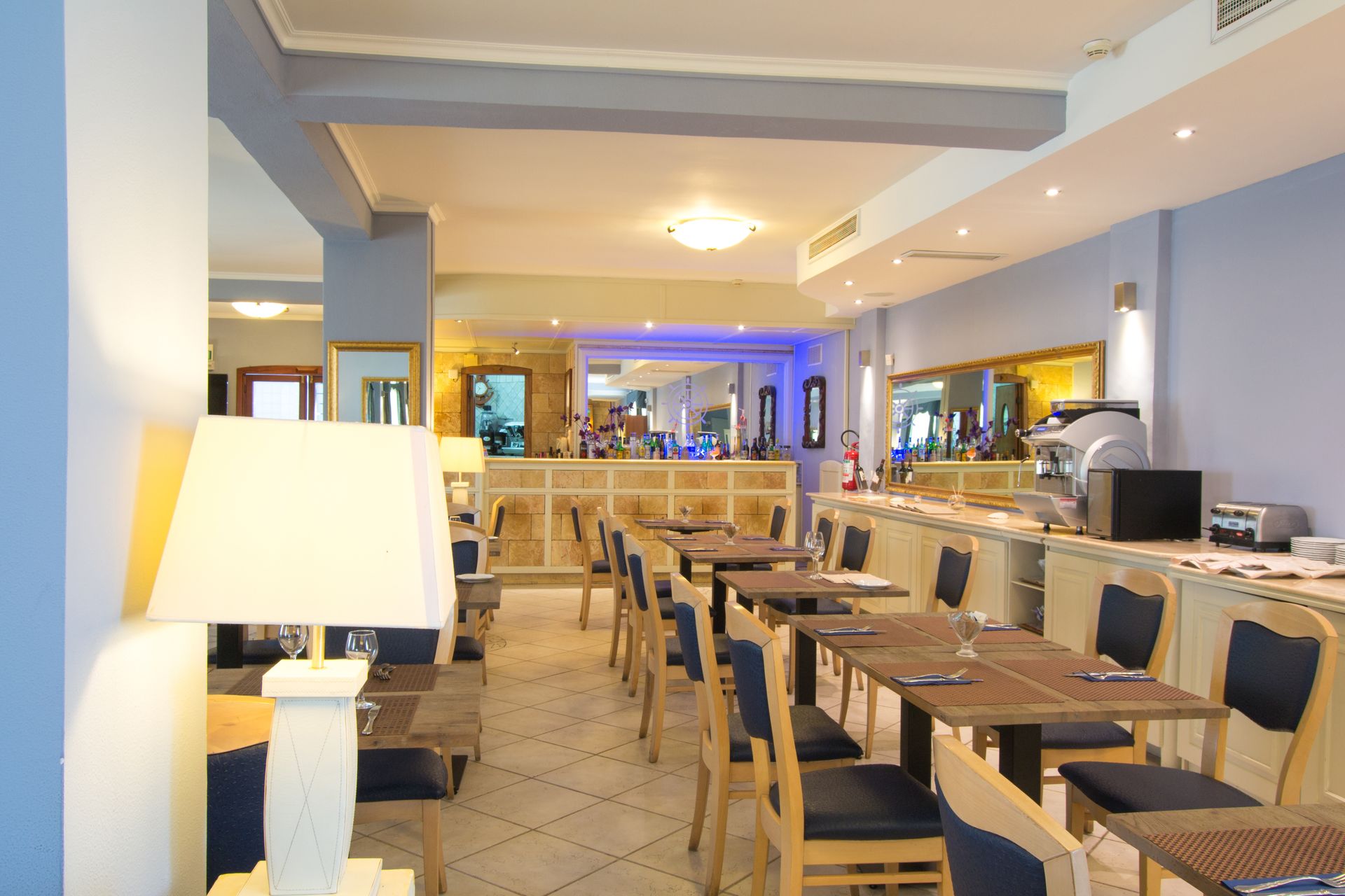 Interno di una sala da pranzo con tavoli e sedie, un buffet e illuminazione dall'alto. Combinazione di colori blu e bianco.