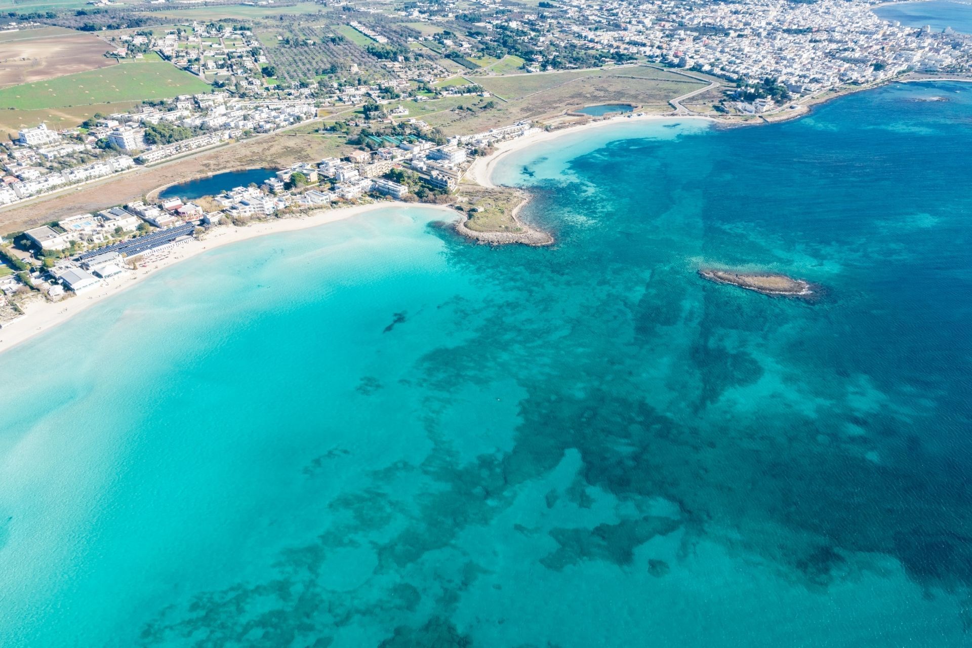 Vista aerea di un mare turchese e di una spiaggia sabbiosa, con una città costiera sullo sfondo.