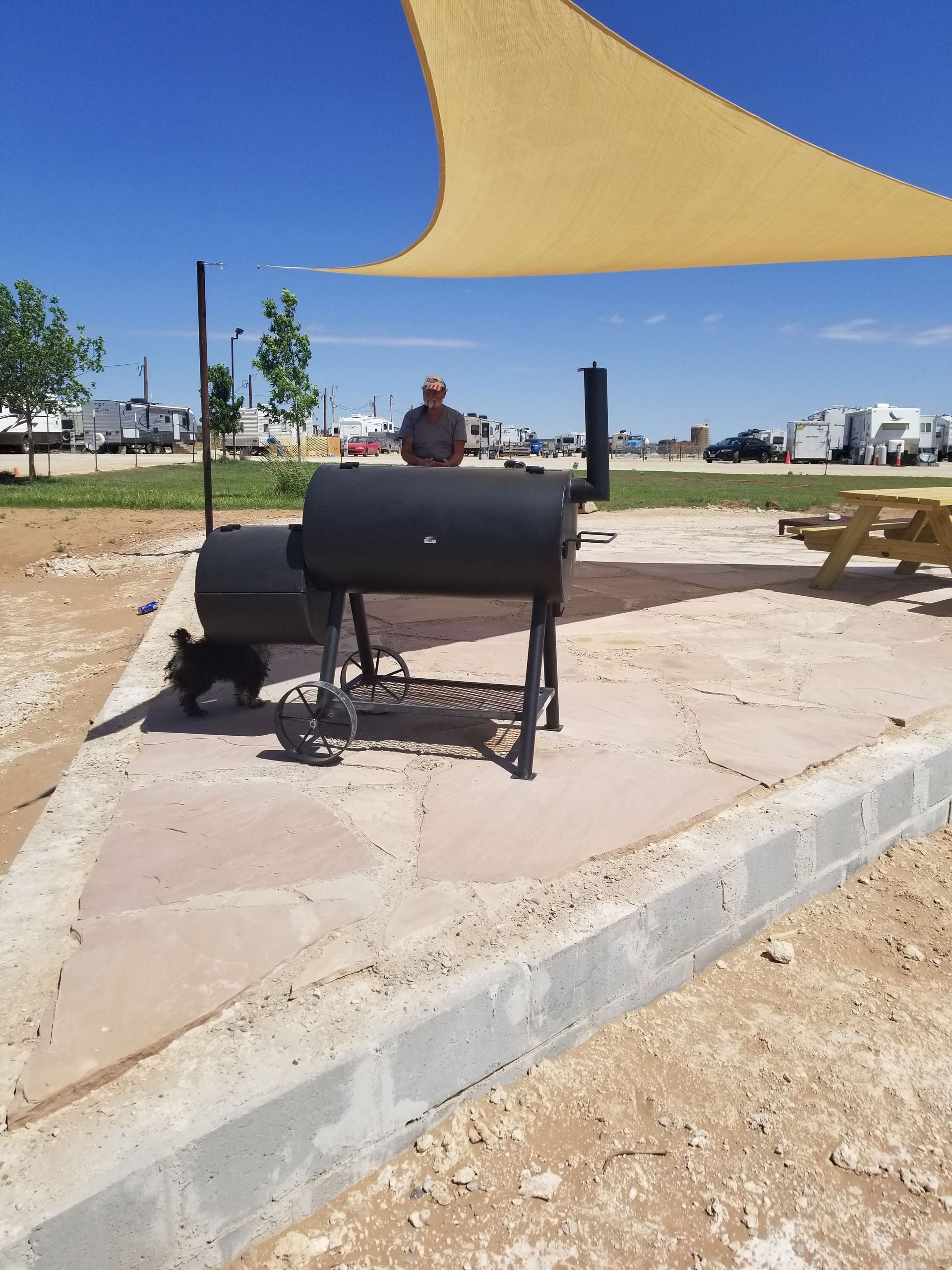 A person stands behind a large black charcoal grill on a stone patio under a tan sun shade in an RV park.