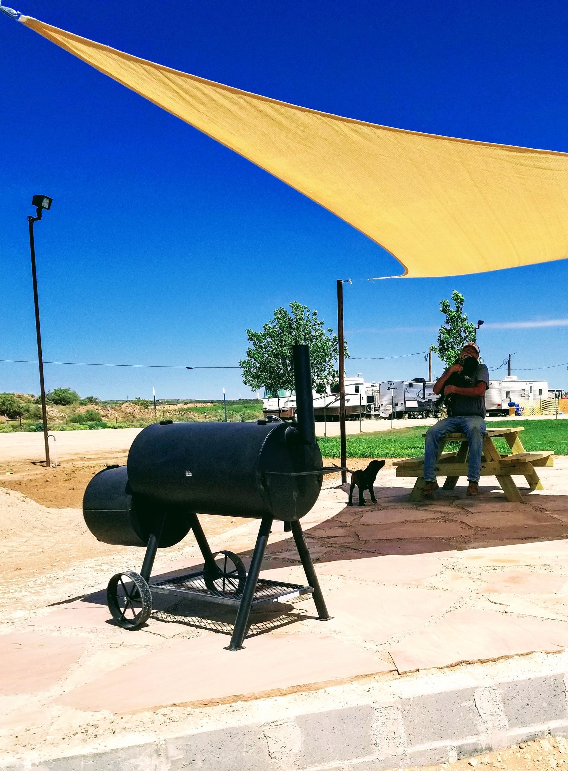 A person sits at a picnic table near a black metal smoker grill under a large tan shade sail on a sunny day.
