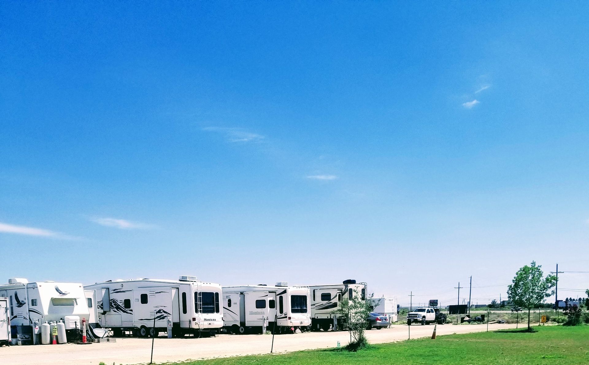 A line of white recreational vehicles parked on a gravel lot under a clear, bright blue sky.