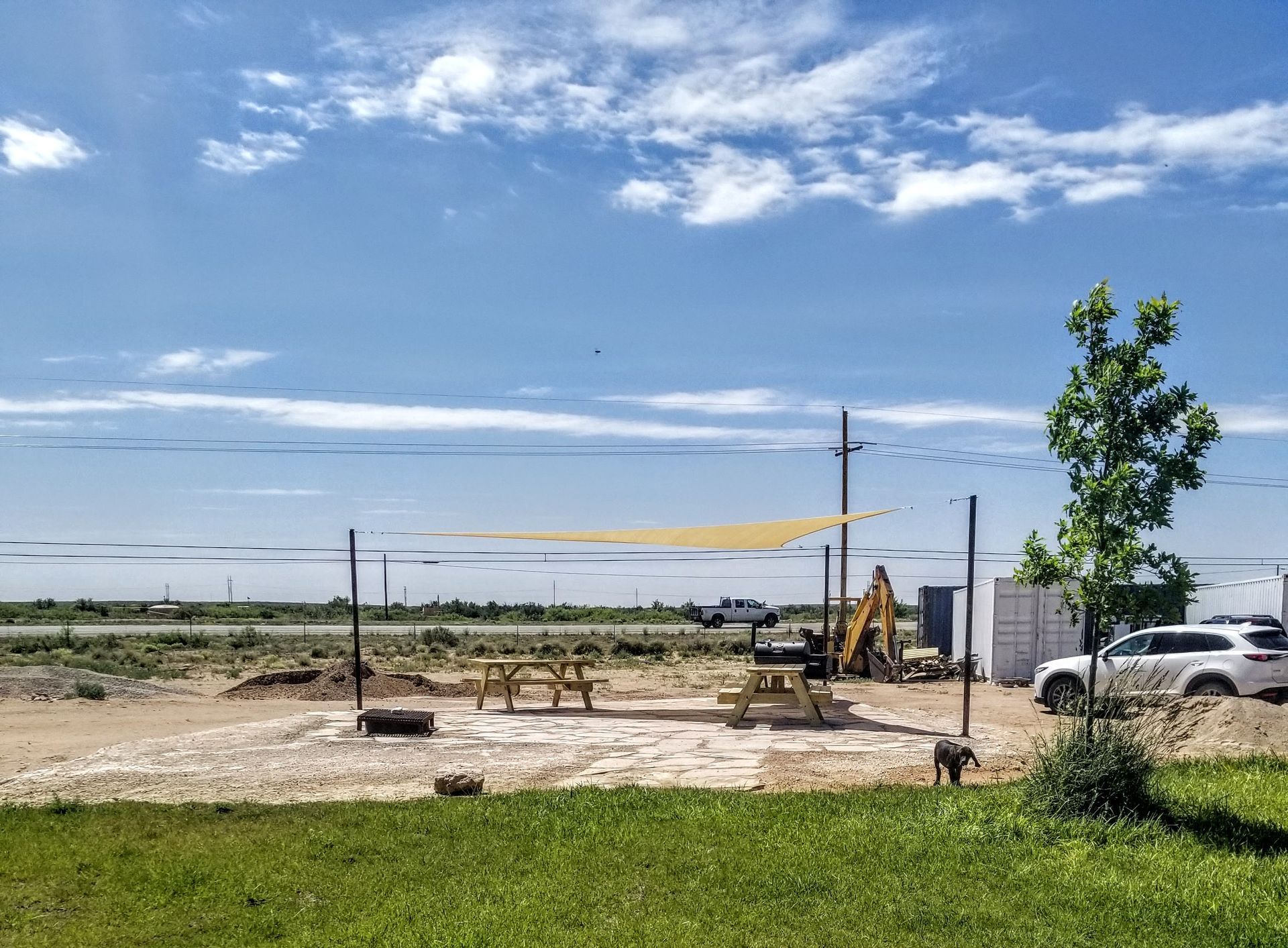 An outdoor area with two picnic tables under a shade sail, set on a gravel lot next to a grassy field and a white vehicle.