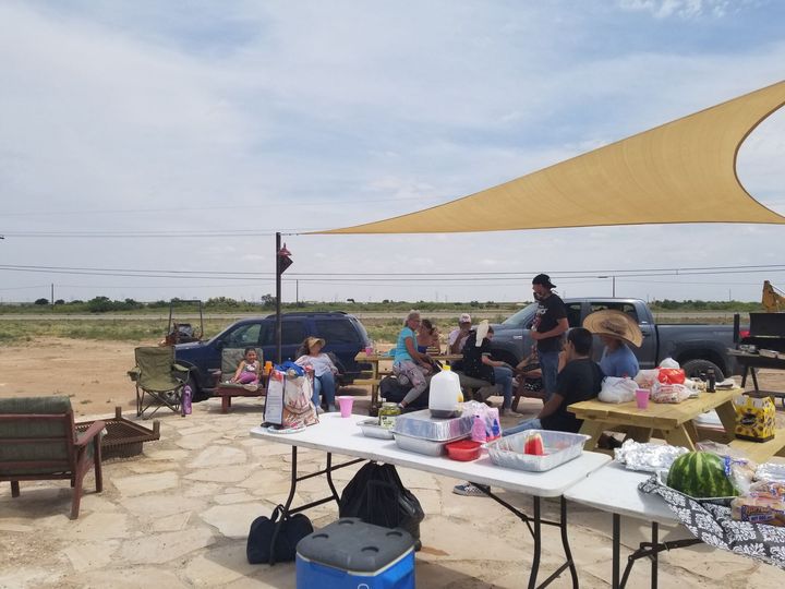 A group of people gather for an outdoor social event under a tan shade sail, with tables, seating, and vehicles nearby.