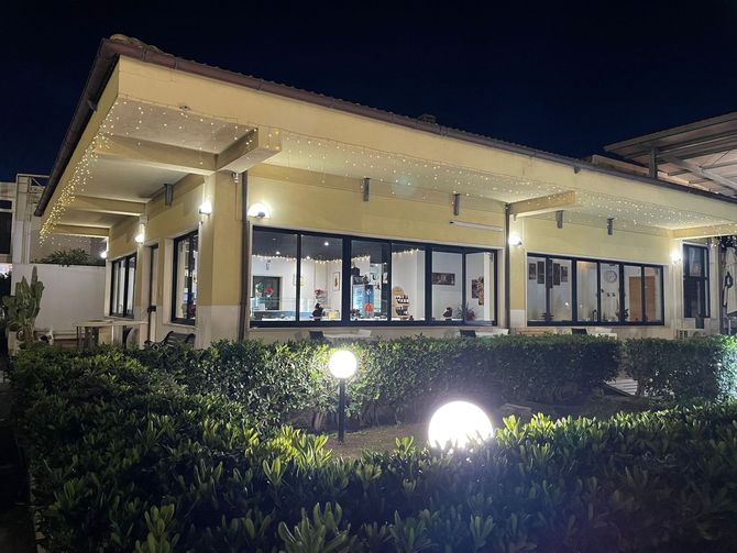 Veduta notturna di un moderno edificio con facciata in vetro, finestre illuminate, siepi e luci da giardino.