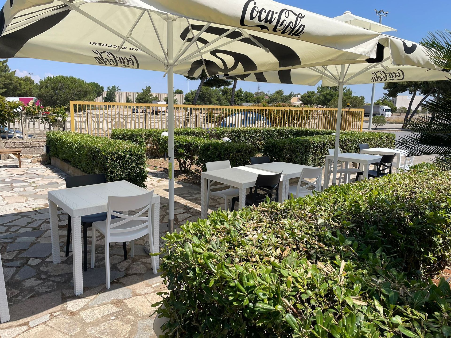 Tavolini da caffè all'aperto con ombrelloni bianchi, tavoli e sedie accanto a siepi ben curate e un patio in pietra.