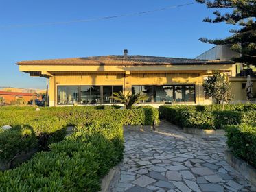 Casa soleggiata con vialetto in pietra, siepi e grandi finestre sotto un cielo azzurro limpido.