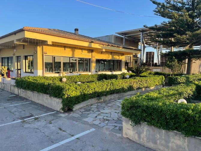 Edificio giallo luminoso con ampie finestre, siepi curate e un'area parcheggio pavimentata.