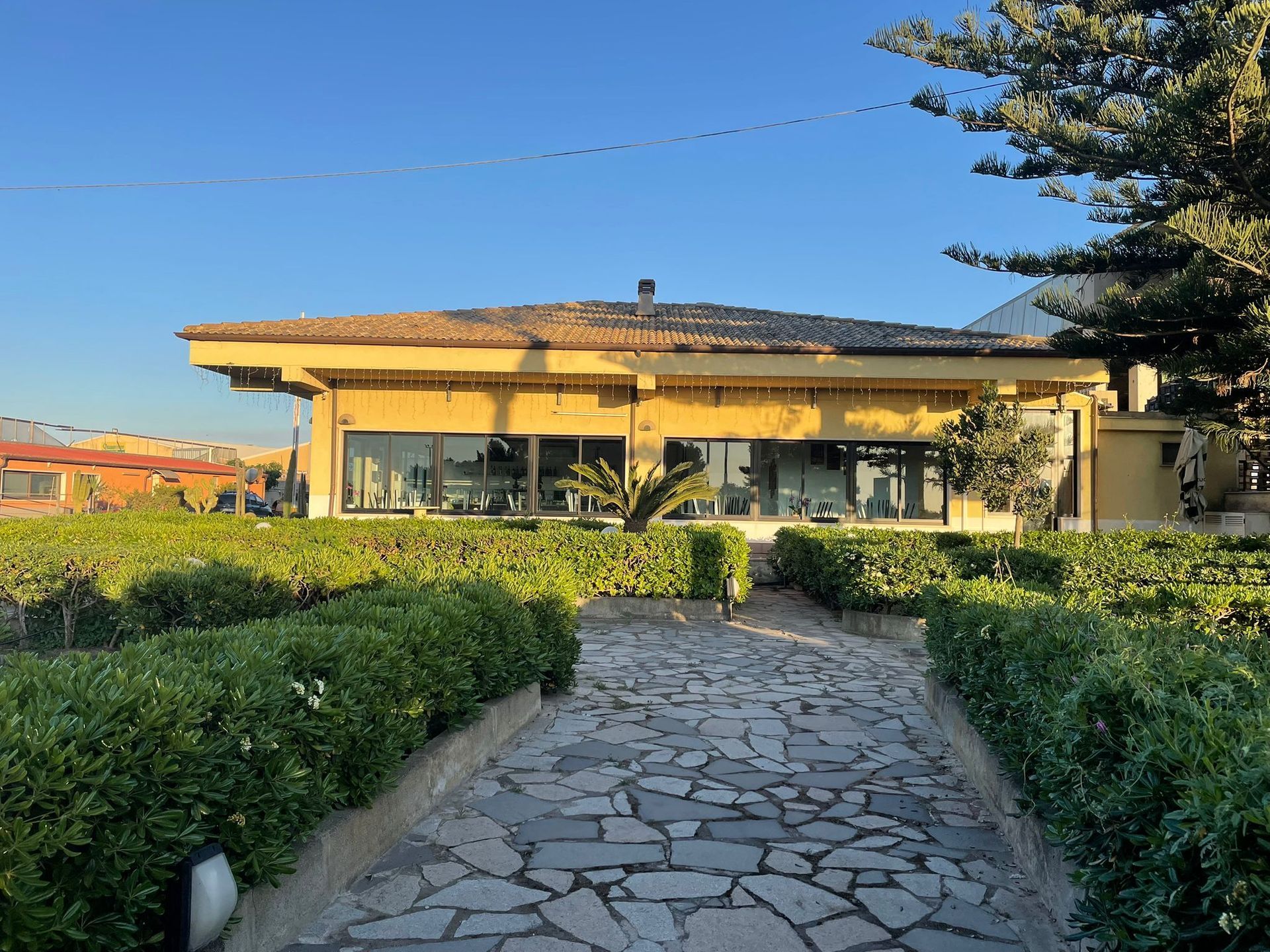 Un sentiero lastricato conduce a una casa moderna con ampie finestre, incorniciata da siepi verdi sotto un cielo azzurro e limpido.