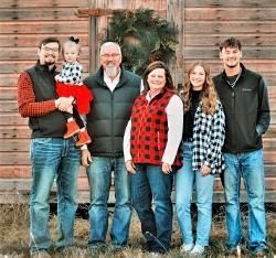 Family poses in front of a wooden shed with a Christmas wreath.