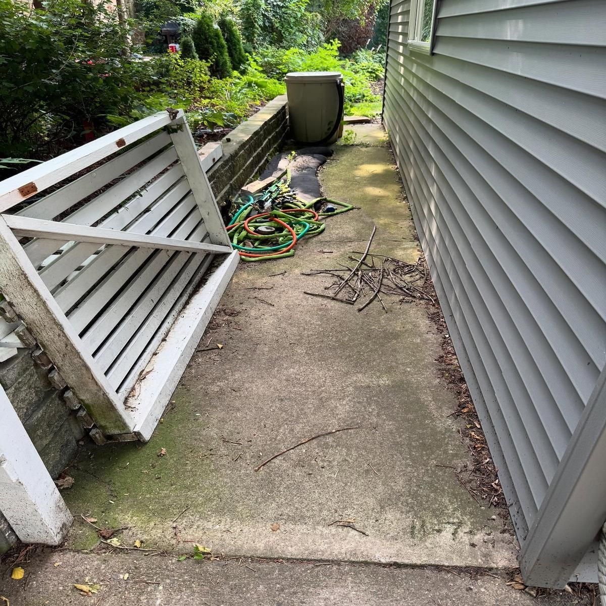 Narrow concrete side yard with a fallen white fence panel, garden hose, trash bin, and house siding.