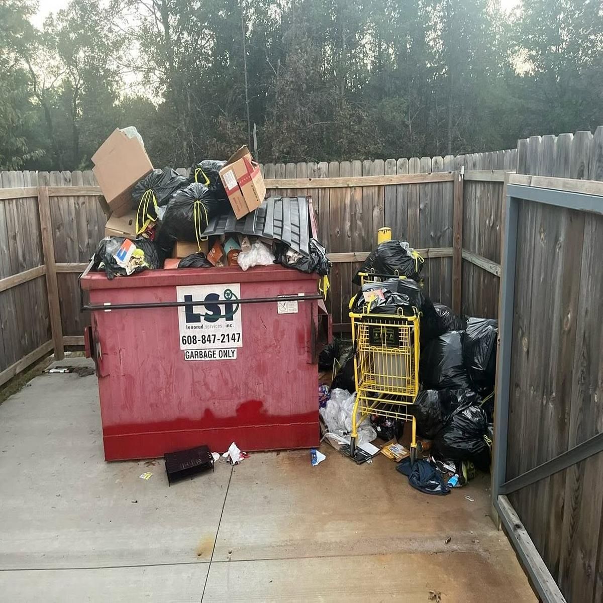 Overflowing red dumpster and piled trash in a fenced alleyway