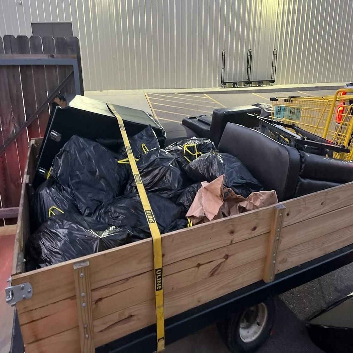 Wooden trailer piled with black trash bags, strapped down with yellow tie-downs in a parking lot.