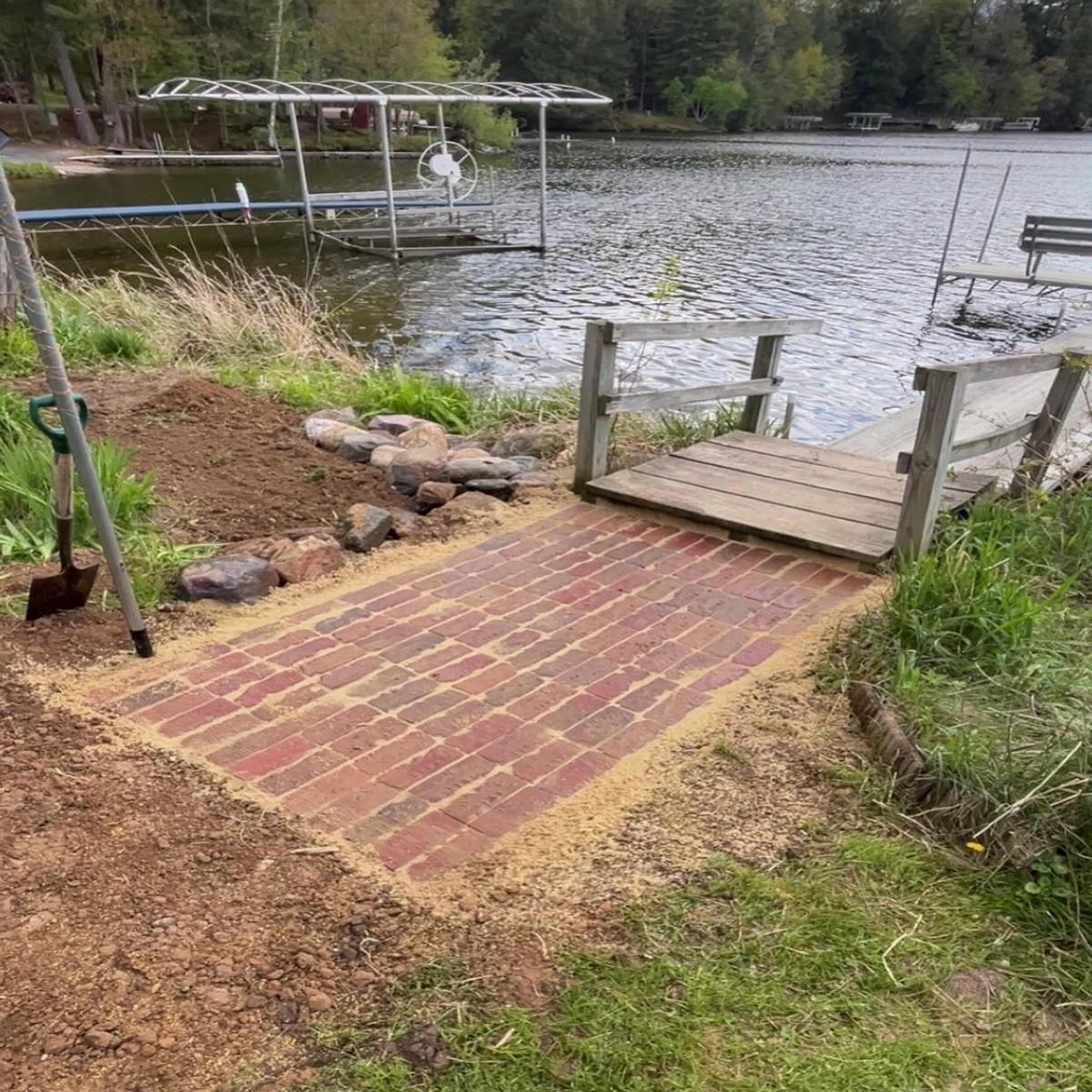 Brick path to a small wooden dock by a lake, with grass, rocks, and trees along the shore