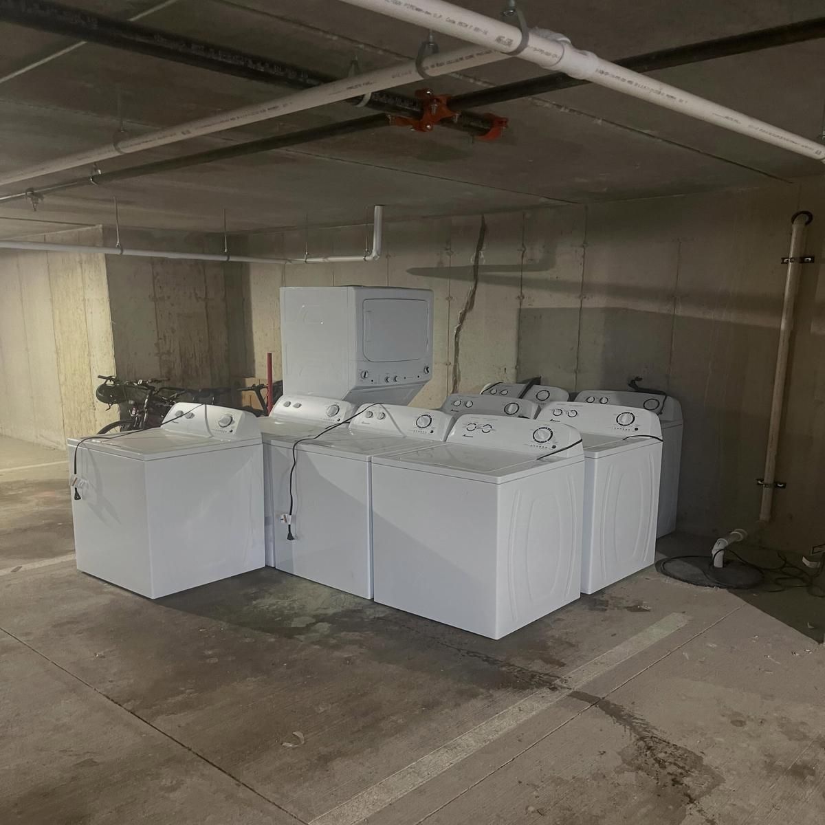 Laundry machines in a concrete basement utility room with exposed pipes and a wall-mounted control box